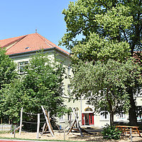 Außengelände: Das Schulgebäude von Montessori Kempten mit Blick über den grünen Schulhof auf die Eingangstüre. 